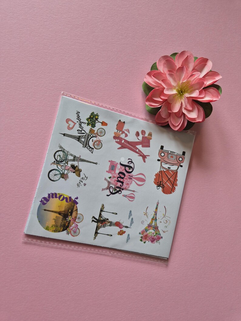 May include: A sheet of Paris-themed stickers featuring the Eiffel Tower, bicycles, and other Parisian imagery. The stickers are in a clear plastic sleeve, with a pink flower in the upper right corner. The background is a solid pink.