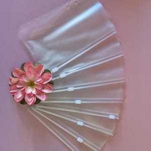May include: Clear plastic zip pouches with white zips are fanned out on a pink surface. A pink and green artificial flower is placed near the pouches. The pouches have holes along the top edge.