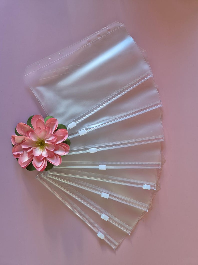 May include: Clear plastic zip pouches with white zips are fanned out on a pink surface. A pink and green artificial flower is placed near the pouches. The pouches have holes along the top edge.