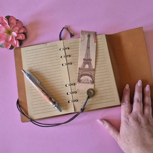 Peut inclure: Un journal en cuir ouvert avec des pages lign&eacute;es, un stylo et un marque-page repr&eacute;sentant la Tour Eiffel. Une fleur rose et une main sont &eacute;galement visibles. Le journal est ouvert sur une surface rose.