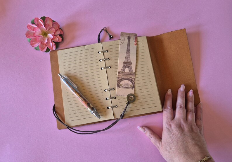 Peut inclure: Un journal en cuir ouvert avec des pages lign&eacute;es, un stylo et un marque-page repr&eacute;sentant la Tour Eiffel. Une fleur rose et une main sont &eacute;galement visibles. Le journal est ouvert sur une surface rose.