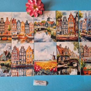 Op de afbeelding: Negen aquarelprints met sc&egrave;nes van Amsterdam, Holland, met grachten, windmolens en kleurrijke gebouwen. De prints zijn gerangschikt in een raster, met een roze bloem bovenaan. Een klein label onderaan luidt "Amsterdam/Holland".