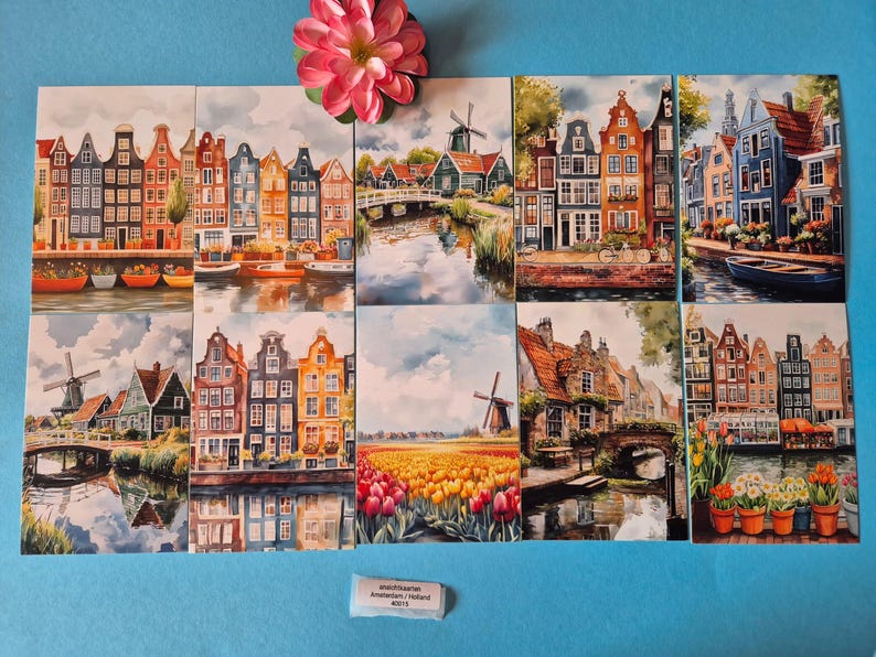 Op de afbeelding: Negen aquarelprints met sc&egrave;nes van Amsterdam, Holland, met grachten, windmolens en kleurrijke gebouwen. De prints zijn gerangschikt in een raster, met een roze bloem bovenaan. Een klein label onderaan luidt "Amsterdam/Holland".