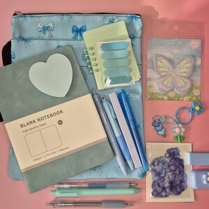 K&ouml;nnte beinhalten: Ein Schreibwaren-Set bestehend aus einer hellblauen Tasche mit Schleifenmuster, einem salbeigr&uuml;nen A5-Notizbuch mit herzf&ouml;rmigem Haftnotizzettel, einem kleinen Spiralblock, einem Schmetterlings-Notizblock, drei Stiften, zwei Schl&uuml;sselanh&auml;ngern, Blumenaufklebern und einem Korrekturroller.