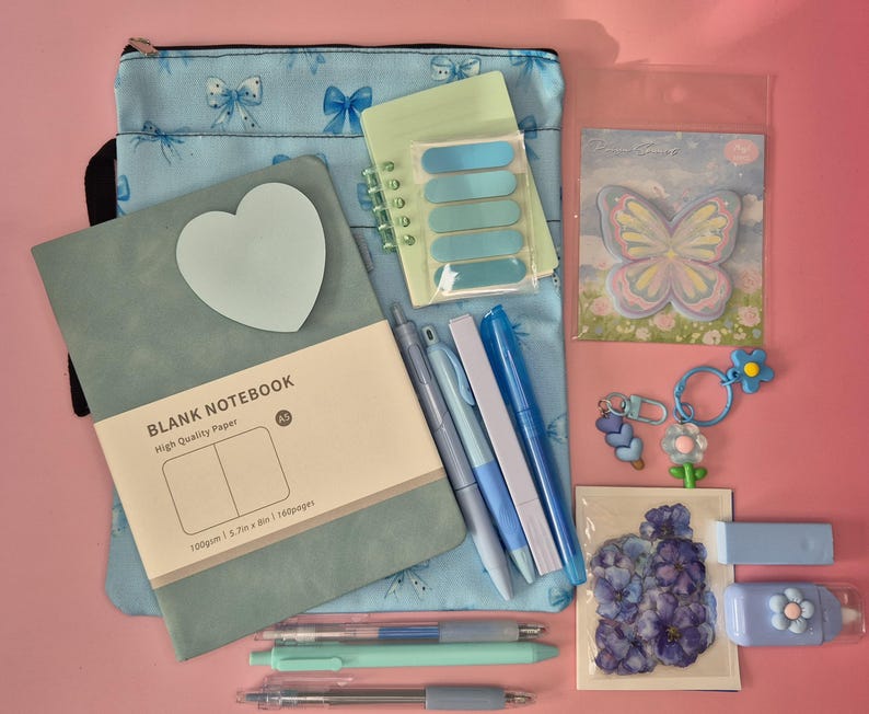 K&ouml;nnte beinhalten: Ein Schreibwaren-Set bestehend aus einer hellblauen Tasche mit Schleifenmuster, einem salbeigr&uuml;nen A5-Notizbuch mit herzf&ouml;rmigem Haftnotizzettel, einem kleinen Spiralblock, einem Schmetterlings-Notizblock, drei Stiften, zwei Schl&uuml;sselanh&auml;ngern, Blumenaufklebern und einem Korrekturroller.