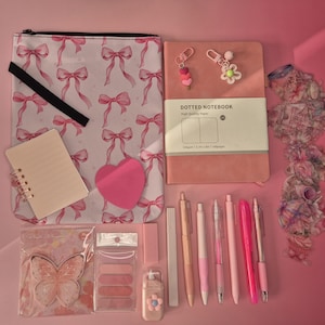 May include: A collection of pink stationery items including a bow-patterned pouch, a dotted notebook with two charms, a spiral notepad, a heart-shaped sticky note, a butterfly-shaped notepad, various pens, markers, and a small flower-adorned correction tape.