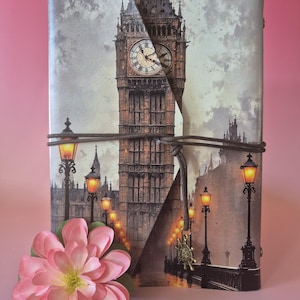 May include: A journal with a Big Ben design, featuring the clock tower and street lamps. A brown cord wraps around the journal. A pink flower is in the foreground. The background is a soft pink.