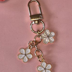 Puede incluir: Un llavero dorado con un cierre y tres colgantes de flores blancas. Cada flor tiene un contorno dorado y un centro blanco. El llavero est&aacute; sobre un fondo rosa.