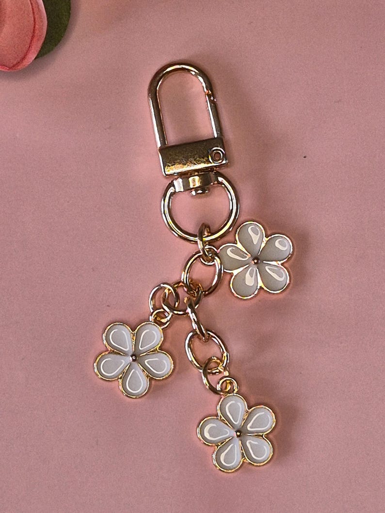 Puede incluir: Un llavero dorado con un cierre y tres colgantes de flores blancas. Cada flor tiene un contorno dorado y un centro blanco. El llavero est&aacute; sobre un fondo rosa.