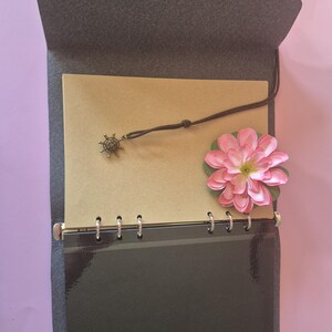 May include: Open gray felt journal with a brown paper interior. A small, decorative charm hangs from a leather cord. A pink flower with green leaves rests on the right side. The journal is open, revealing the inside pages.