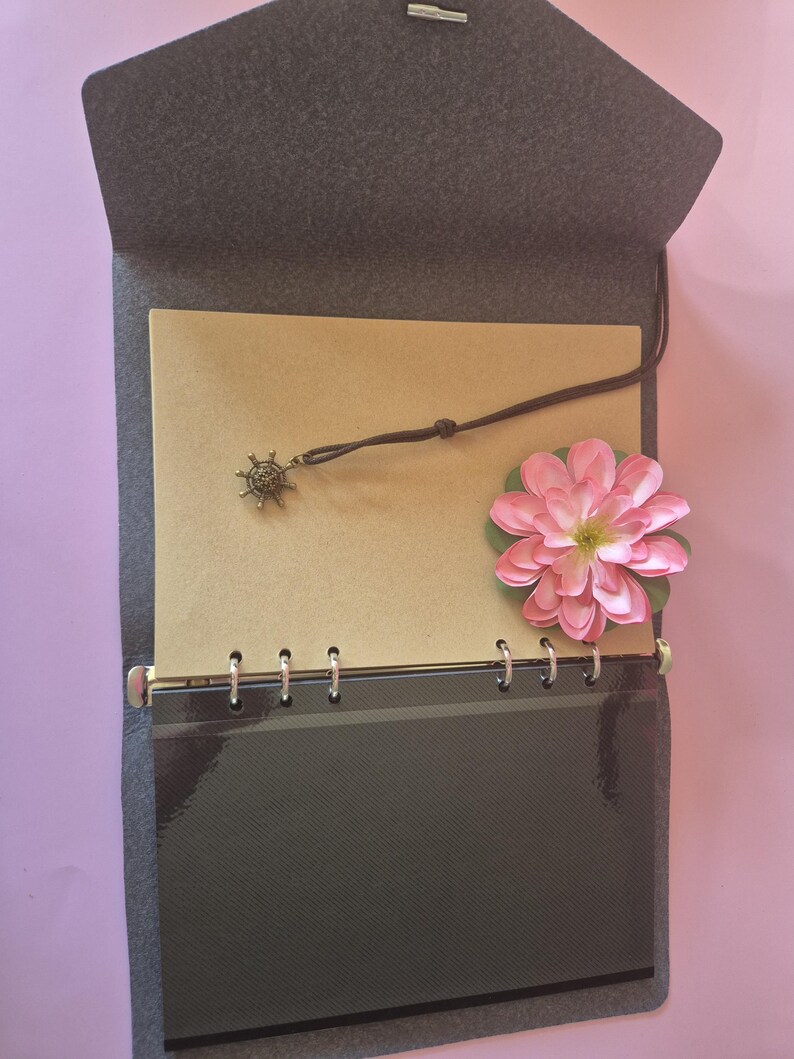 May include: Open gray felt journal with a brown paper interior. A small, decorative charm hangs from a leather cord. A pink flower with green leaves rests on the right side. The journal is open, revealing the inside pages.