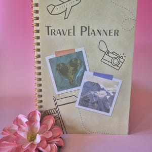 Op de afbeelding: Een spiraalgebonden reisplanner met de tekst "Travel Planner" en illustraties van een vliegtuig, camera en vlag. Twee foto's in polaroidstijl zijn op de omslag geplakt. Een roze bloem ligt op de voorgrond.