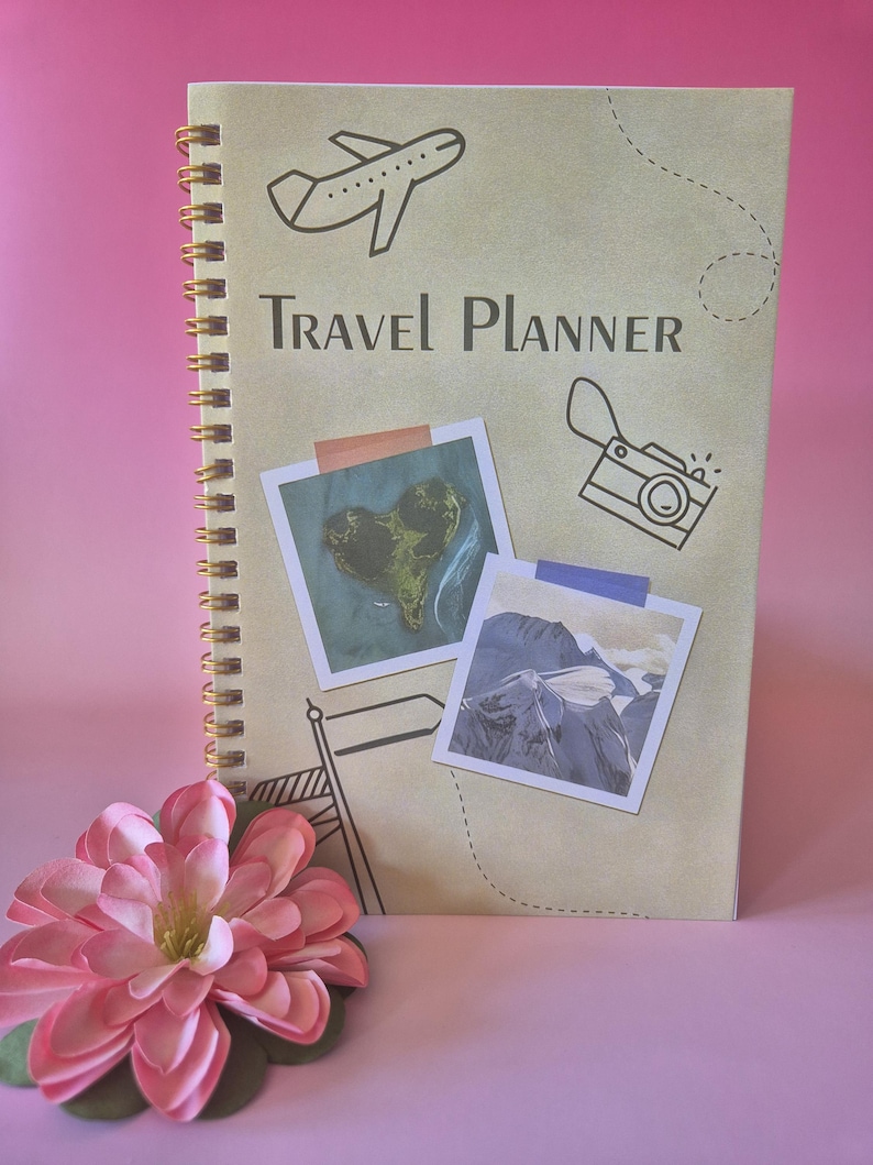 Op de afbeelding: Een spiraalgebonden reisplanner met de tekst "Travel Planner" en illustraties van een vliegtuig, camera en vlag. Twee foto's in polaroidstijl zijn op de omslag geplakt. Een roze bloem ligt op de voorgrond.