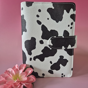 Op de afbeelding: Een koeienprint planner in wit en zwart, met een magnetische sluiting. De rechthoekige planner staat naast een roze bloem. De planner is gemaakt van een textuurrijk materiaal.