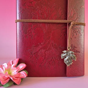May include: A burgundy leather journal with an embossed tree and flying birds design. A brown leather cord wraps around the journal, and a metal leaf charm hangs from the side. A pink flower rests on the lower left.