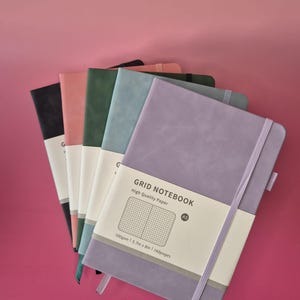 May include: A stack of five A5 grid notebooks with matte covers in purple, blue, green, pink, and black. Each notebook features an elastic closure band and a white paper belly band printed with GRID NOTEBOOK, High Quality Paper, 100gsm, 14.5cm x 20.3cm, 160pages.