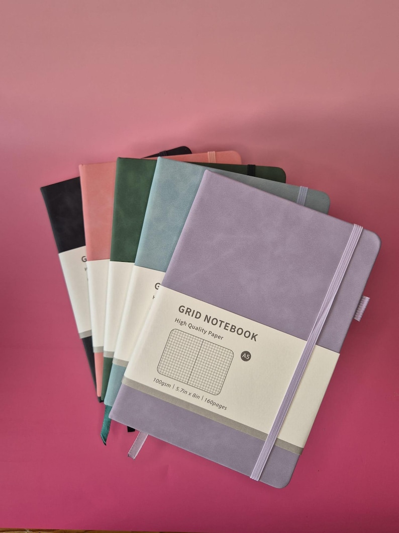 May include: A stack of five A5 grid notebooks with matte covers in purple, blue, green, pink, and black. Each notebook features an elastic closure band and a white paper belly band printed with GRID NOTEBOOK, High Quality Paper, 100gsm, 14.5cm x 20.3cm, 160pages.