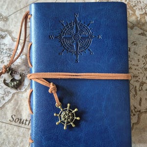 May include: A blue leather journal with a compass rose design embossed on the cover. The journal is secured with a brown leather strap and features a decorative anchor and ship wheel charm.