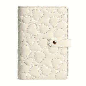 May include: Cream-colored planner with a quilted heart pattern. The cover features a stitched heart design and a matching strap with a gold-colored snap closure. The planner is closed and ready for use.