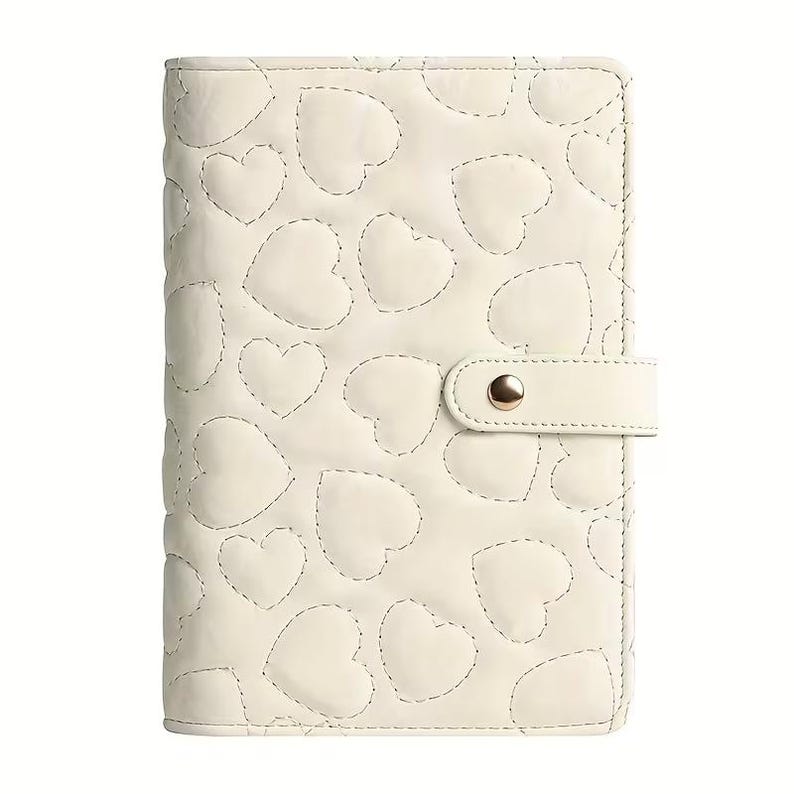 May include: Cream-colored planner with a quilted heart pattern. The cover features a stitched heart design and a matching strap with a gold-colored snap closure. The planner is closed and ready for use.