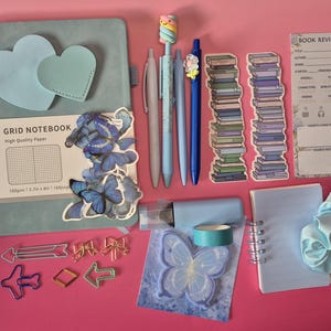 May include: A light blue grid notebook with heart-shaped accents, blue butterfly stickers, four pens, two book stack bookmarks, a book review card, arrow and bow paperclips, a light blue tape roll, a butterfly sticky note, and a light blue scrunchie.