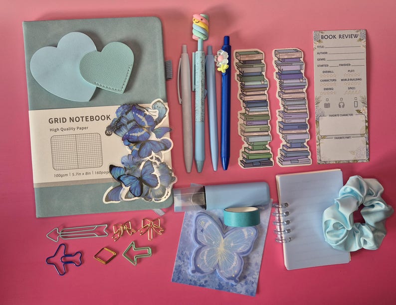 May include: A light blue grid notebook with heart-shaped accents, blue butterfly stickers, four pens, two book stack bookmarks, a book review card, arrow and bow paperclips, a light blue tape roll, a butterfly sticky note, and a light blue scrunchie.