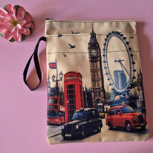 Op de afbeelding: Een beige etui met ritssluiting met een Londense stadsgezicht, waaronder de Big Ben, de London Eye, een rode telefooncel en dubbeldekkersbussen. Een zwart handvat is bevestigd. Een roze bloem staat in de linkerbovenhoek.