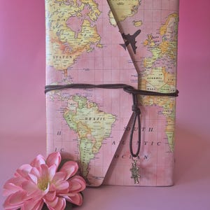 May include: A pink journal with a world map design, secured with a brown cord and a small charm. A decorative pink flower is in the foreground. The journal features a stylized airplane graphic.