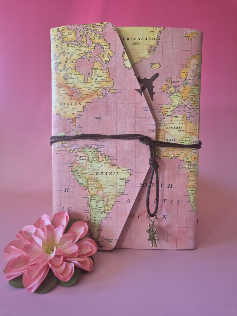 May include: A pink journal with a world map design, secured with a brown cord and a small charm. A decorative pink flower is in the foreground. The journal features a stylized airplane graphic.
