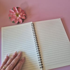 May include: An open, lined notebook with a black spiral binding rests on a pink surface. A pink and green artificial flower is placed near the notebook. The notebook's pages are blank and ready for writing.