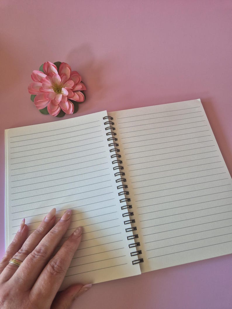 May include: An open, lined notebook with a black spiral binding rests on a pink surface. A pink and green artificial flower is placed near the notebook. The notebook's pages are blank and ready for writing.
