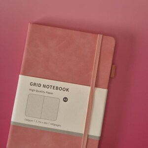 May include: A dusty rose A5 grid notebook with a vertical elastic closure and a pen loop. A white paper band wraps around the cover, featuring the text GRID NOTEBOOK, High Quality Paper, A5, 100gsm, 14.5cm x 20.3cm, and 160pages.