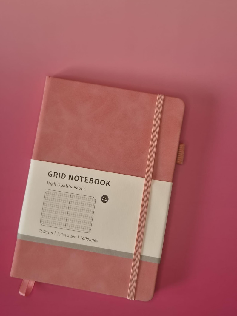 May include: A dusty rose A5 grid notebook with a vertical elastic closure and a pen loop. A white paper band wraps around the cover, featuring the text GRID NOTEBOOK, High Quality Paper, A5, 100gsm, 14.5cm x 20.3cm, and 160pages.