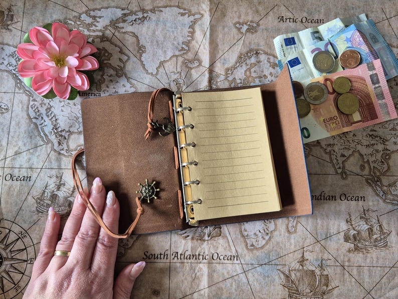 May include: Open brown leather journal with lined pages, a decorative charm, and a leather tie. A pink flower and money are also visible on a map background. The journal is open, revealing blank pages.