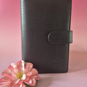 Pu&ograve; includere: Diario marrone scuro con una texture simile alla pelle e una chiusura con fibbia. Un fiore rosa e verde in primo piano. Il diario presenta un emblema in rilievo nell'angolo in basso a destra.