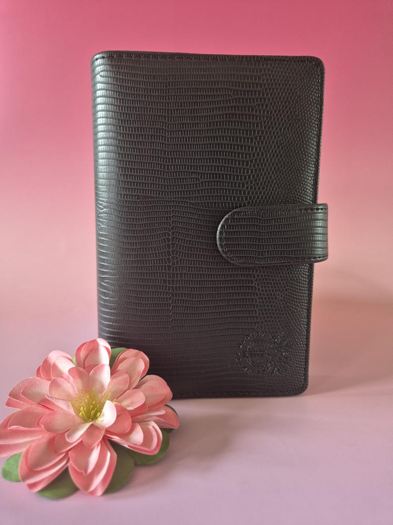 Pu&ograve; includere: Diario marrone scuro con una texture simile alla pelle e una chiusura con fibbia. Un fiore rosa e verde in primo piano. Il diario presenta un emblema in rilievo nell'angolo in basso a destra.