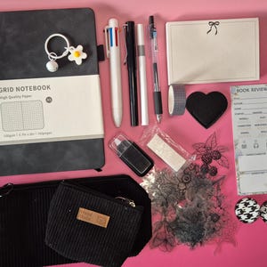 Puede incluir: Un set de papeler&iacute;a que incluye un cuaderno de cuadr&iacute;cula A5 gris oscuro con un llavero de flor, un bol&iacute;grafo multicolor, bol&iacute;grafos negro y blanco, un bloc de notas con lazo, un parche de coraz&oacute;n, una tarjeta de rese&ntilde;a de libros y sellos.
