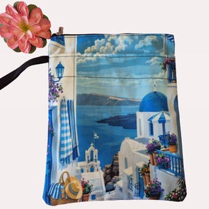 Puede incluir: Una bolsa cuadrada de tela azul y blanca con cierre de cremallera. La bolsa presenta un estampado esc&eacute;nico de un pueblo griego con edificios blancos, c&uacute;pulas azules y el mar. Una flor rosa y una correa negra est&aacute;n unidas a la bolsa.