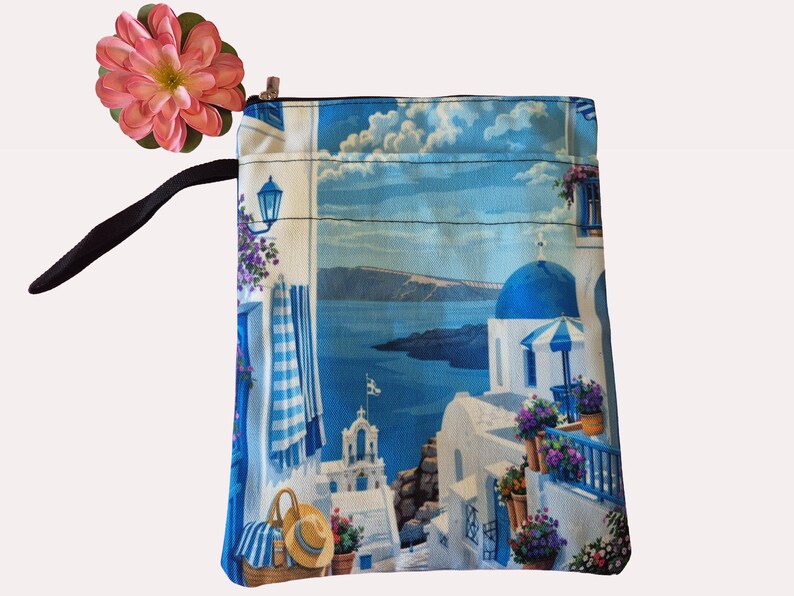 Puede incluir: Una bolsa cuadrada de tela azul y blanca con cierre de cremallera. La bolsa presenta un estampado esc&eacute;nico de un pueblo griego con edificios blancos, c&uacute;pulas azules y el mar. Una flor rosa y una correa negra est&aacute;n unidas a la bolsa.
