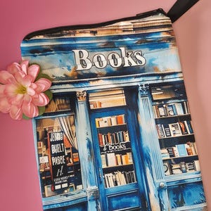 May include: A rectangular, blue and white zippered pouch with a bookstore design. The pouch features the word "Books" at the top and a detailed illustration of a bookstore facade. A pink flower is in the corner.