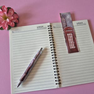 Op de afbeelding: Een open spiraalgebonden notitieboek met gelinieerde pagina's, een pen, een bladwijzer en een roze bloem op een roze achtergrond. De bladwijzer toont een rode telefooncel. De pen heeft een transparante schacht met een decoratief ontwerp. De bloem is roze met groene bladeren.