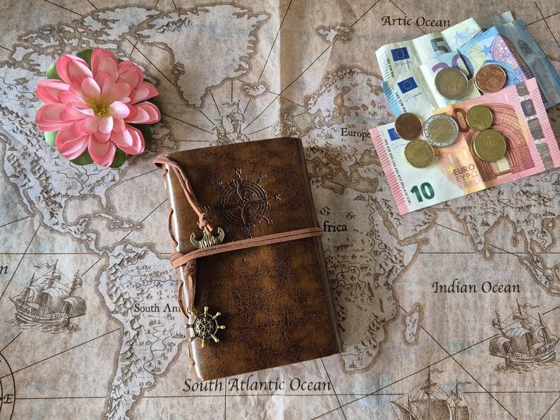 May include: A brown leather journal with a compass rose design and a decorative charm, resting on a vintage world map. A pink flower and Euro currency are also present, evoking a travel theme.
