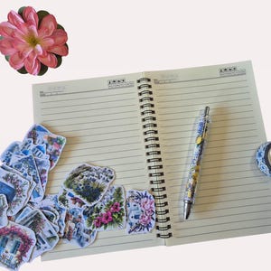 Puede incluir: Un cuaderno espiral abierto con p&aacute;ginas rayadas, un bol&iacute;grafo, pegatinas decorativas, un rollo de cinta washi y una flor rosa. El bol&iacute;grafo tiene un cuerpo transparente con un dise&ntilde;o floral. Las pegatinas representan escenas con flores y edificios.