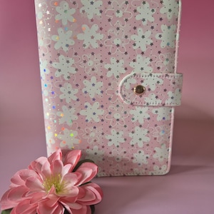 K&ouml;nnte beinhalten: Ein rosa Planer mit floralem Muster und einem Schnappverschluss. Der Einband zeigt wei&szlig;e Blumen und Sterne mit schillernden Akzenten. Eine rosa Blume mit gr&uuml;nen Bl&auml;ttern liegt vor dem Planer.