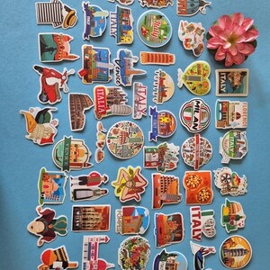 May include: A collection of colorful Italy-themed stickers and magnets, featuring iconic landmarks, food, and cultural symbols. Designs include the Leaning Tower of Pisa, pizza, and the Colosseum. A pink flower is also present.