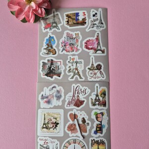 May include: A collection of Paris-themed stickers featuring the Eiffel Tower, bicycles, and floral designs. The stickers are in various shapes and sizes, with a pink flower in the upper left corner. The stickers are on a gray background.
