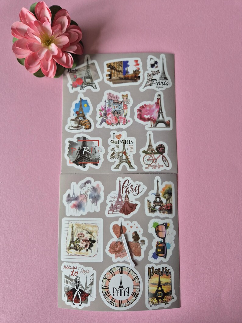 May include: A collection of Paris-themed stickers featuring the Eiffel Tower, bicycles, and floral designs. The stickers are in various shapes and sizes, with a pink flower in the upper left corner. The stickers are on a gray background.