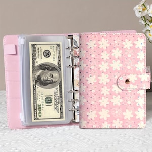 K&ouml;nnte beinhalten: Rosa Budget-Ordner mit floralem Muster und Druckknopfverschluss. Der Ordner ist ge&ouml;ffnet und zeigt eine transparente Tasche mit einem 100-Dollar-Schein. Der Ordner hat einen Ringmechanismus zum Organisieren von Eins&auml;tzen.