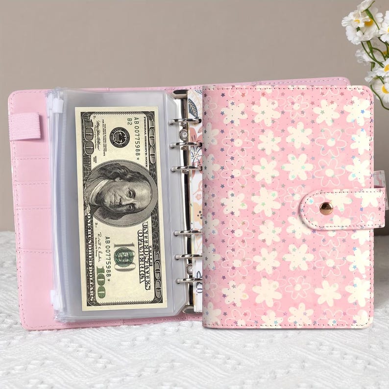 K&ouml;nnte beinhalten: Rosa Budget-Ordner mit floralem Muster und Druckknopfverschluss. Der Ordner ist ge&ouml;ffnet und zeigt eine transparente Tasche mit einem 100-Dollar-Schein. Der Ordner hat einen Ringmechanismus zum Organisieren von Eins&auml;tzen.