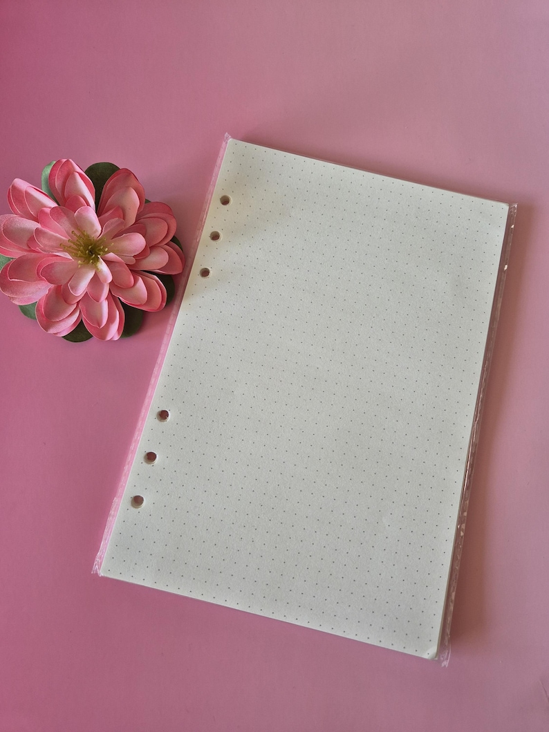 K&ouml;nnte beinhalten: Ein Stapel Punktrasterpapier mit Lochung, verpackt in durchsichtigem Kunststoff. Links vom Papier befindet sich eine rosa-gr&uuml;ne Kunstblume. Der Hintergrund ist einfarbig rosa.